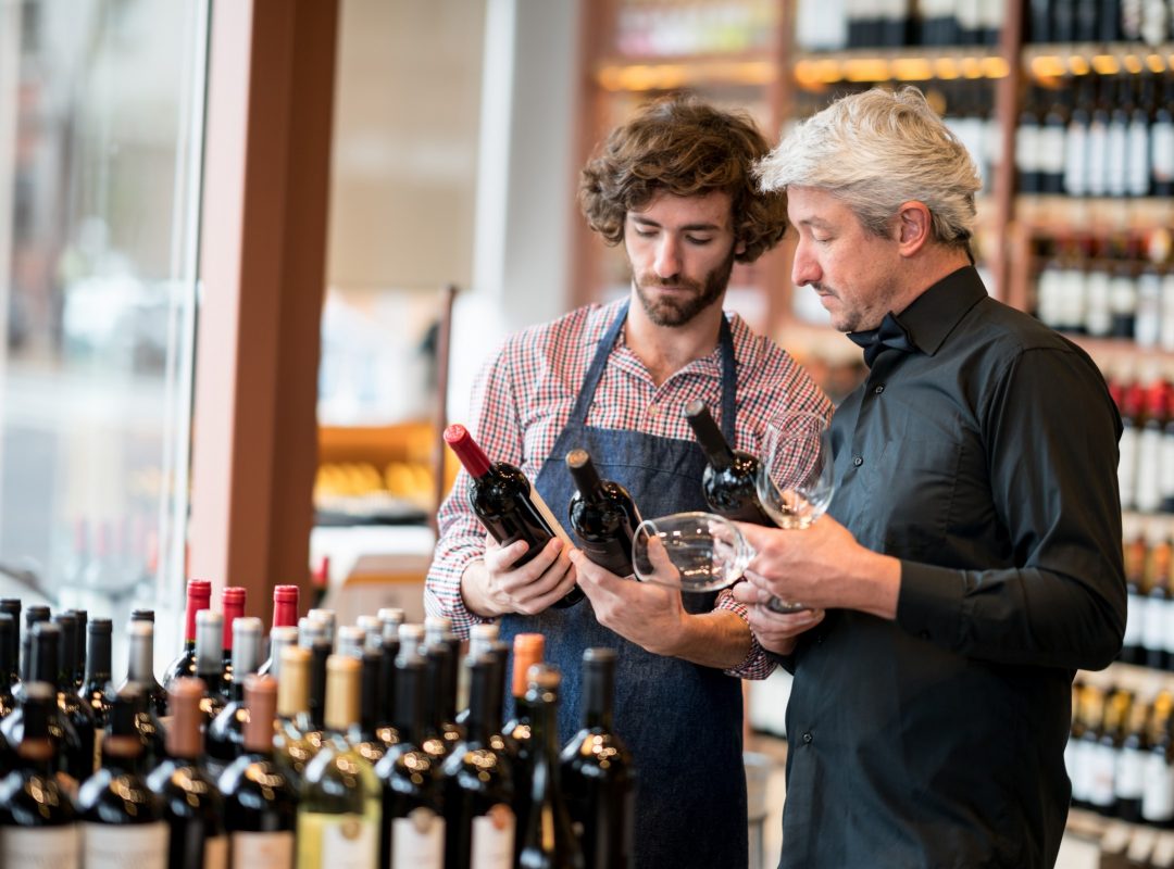 Young sales clerk and business owner looking at bottles of wine while owner holds wine glasses at a winery both looking very serious